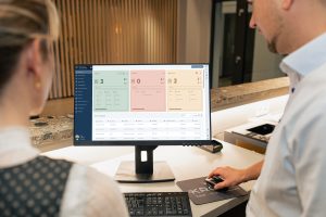 Zwei Hotelmitarbeiter an der Rezeption blicken auf einen Monitor mit dem SoftTec Cloud Hotelsoftware-Dashboard, das Check-ins, Check-outs und aktuelle Hausgäste in farbigen Kacheln anzeigt.